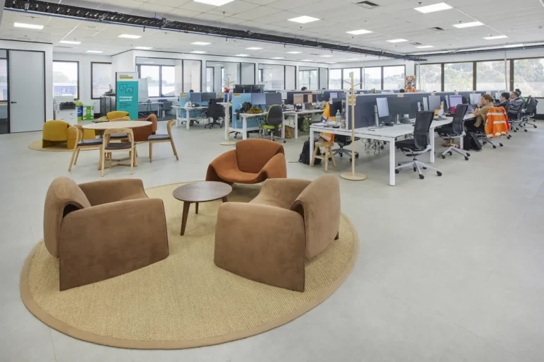 Breakout zone with brown armchairs on a round rug beside open-plan desks, CPB Contractors office fitout, Blackburn North.