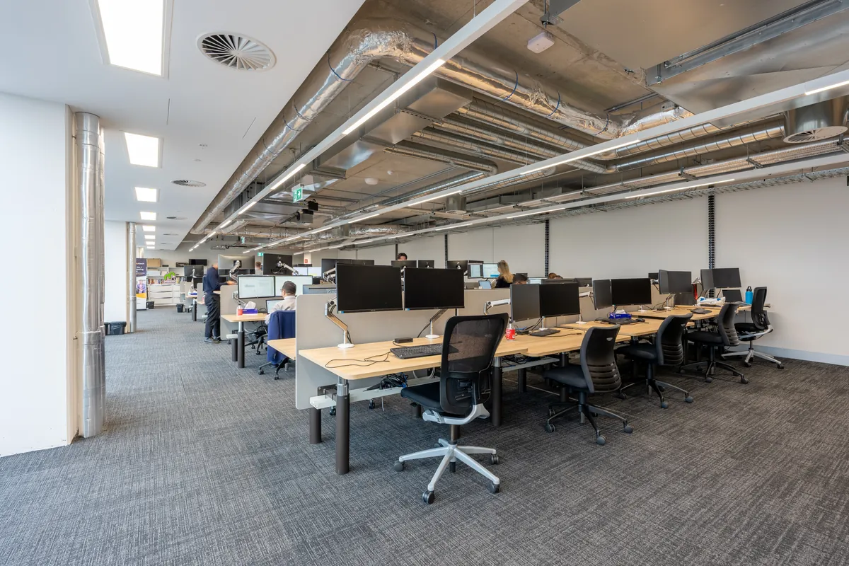 Open-plan workstation zone with staff at timber desks under exposed services at the Empower Wealth office fitout, North Melbourne.