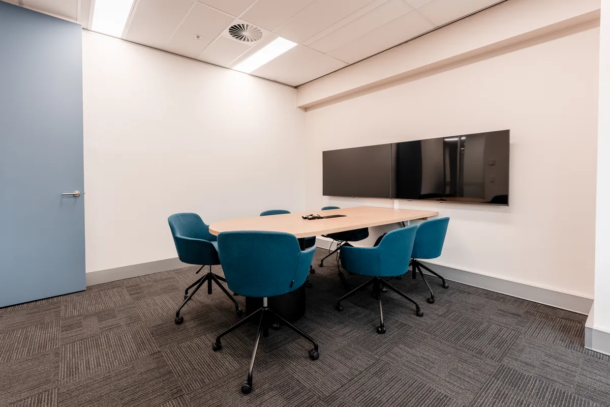 Compact meeting room with timber table, blue chairs and wall-mounted screen at the McLarens office fitout, Melbourne.